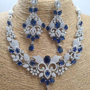 May include: A silver necklace and earrings set with a floral design and blue stones. The necklace has a pendant with a large blue stone.
