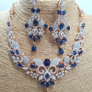 May include: A rose gold necklace and earrings set with a floral design and blue stones. The necklace has a pendant with a teardrop-shaped blue stone.
