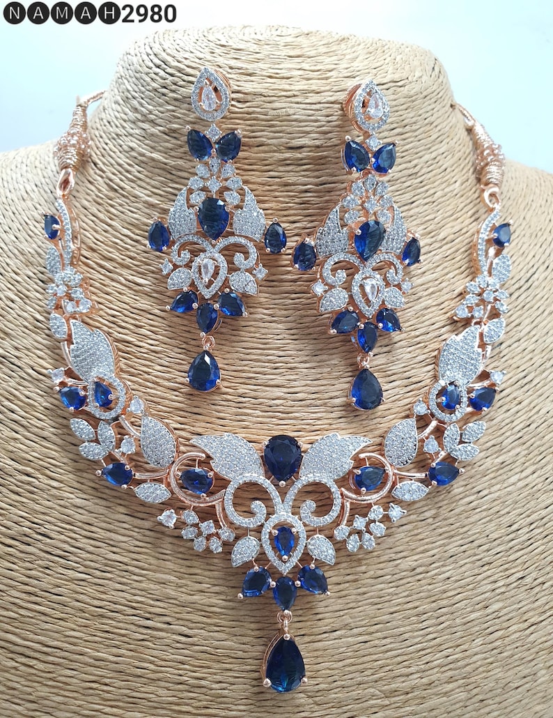 May include: A rose gold necklace and earrings set with a floral design and blue stones. The necklace has a pendant with a teardrop-shaped blue stone.