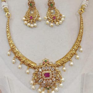 Op de afbeelding: Een gouden ketting met een hanger met een vierkante steen omringd door kleinere stenen. De ketting heeft een halvemaanvorm en is versierd met witte parels. Passende oorbellen met groene en roze stenen zijn ook inbegrepen.