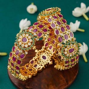 May include: A pair of gold bangles with intricate designs featuring red and green gemstones. The bangles are adorned with small, round white stones.