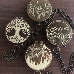 May include: Four brass compass pendants with different nature-themed designs. The pendants feature a tree of life, a mountain scene with a river, a mountain scene with a sun, and a mountain scene with a moon. Each pendant is attached to a brass chain.