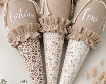 May include: Three cone-shaped fabric gift bags with beige muslin tops, white drawstring closures, and ruffled accents. The left bag features a leopard print, while the center and right bags display delicate brown floral patterns. Names Carlotta and Leni are printed in white.