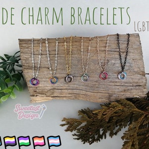 May include: Six pride charm bracelets with different coloured rings on a wooden surface. The bracelets are on chains of silver, gold, rose gold, and black. The text "LGBTQA+" is in the top right corner of the image.  The text "Sweetest Design" is in the bottom left corner of the image.  Six pride flags are at the bottom of the image.
