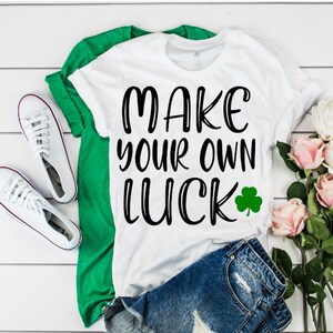 Puede incluir: Camiseta blanca con el texto "MAKE YOUR OWN LUCK" en letras negras y un trébol verde. La camiseta está doblada y se muestra sobre una superficie de madera blanca con un par de zapatillas blancas, una camiseta verde y flores rosas.