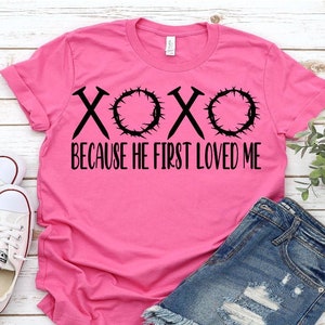 May include: Pink t-shirt with a black graphic that says "XOXO Because He First Loved Me". The graphic includes a crown of thorns and nails.