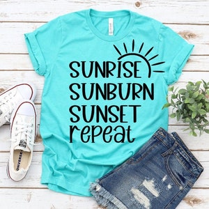 Könnte beinhalten: Ein türkisfarbenes T-Shirt mit schwarzem Text, der "Sunrise Sunburn Sunset Repeat" lautet, und einer Sonnengrafik. Das Shirt liegt auf einer weißen Holzoberfläche mit einem Paar weißen Turnschuhen und einer Jeans-Shorts.