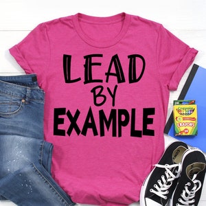 Peut inclure: T-shirt rose avec le texte "LEAD BY EXAMPLE" en lettres noires. Le t-shirt est posé sur une surface blanche avec un jean bleu, une paire de baskets noires et un cahier bleu avec une boîte de crayons.
