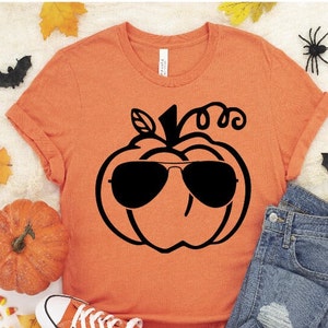 Op de afbeelding: Oranje T-shirt met een zwarte afbeelding van een pompoen met een zonnebril. De pompoen heeft een steel en een gekrulde rank.