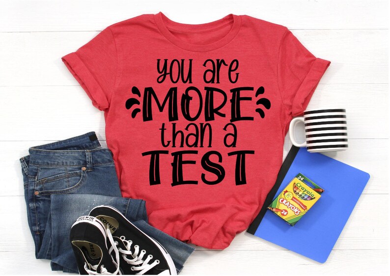 May include: Red t-shirt with black text that reads "You are MORE than a TEST". The shirt is laid flat on a white wooden surface with a pair of blue jeans, a black and white striped mug, a blue binder, and a box of crayons.