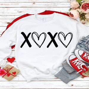 May include: White sweatshirt with black 'XOXO' graphic with hearts. The sweatshirt is laid out on a white wood background with red hearts and a pair of red sneakers.