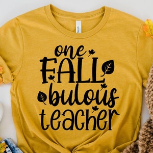 Puede incluir: Una camiseta amarilla mostaza con el texto "one fall-bulous teacher" en letras negras. La camiseta está decorada con pequeños gráficos de hojas.