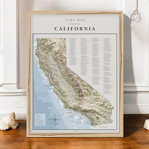 May include: A framed surf map of California, featuring a detailed topographical view of the state. The map includes the text "SURF MAP of the State of CALIFORNIA" and lists surf spots. The frame is made of light-colored wood.