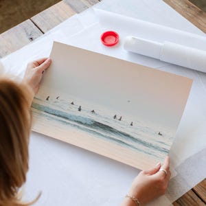A person is holding a framed photograph of a group of people surfing on a beach, with the ocean visible in the background.