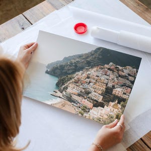 A person is holding a framed photograph of a coastal town, with the ocean visible in the background.