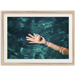 May include: A framed print featuring a hand holding a seashell in clear, turquoise water. The seashell is tan and brown, and the water has ripples. The image is set in a light wood frame.