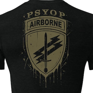 May include: A black short-sleeve t-shirt featuring a graphic on the back. The design includes an olive green shield emblem with the text PSYOP and AIRBORNE, displaying a central sword and two lightning bolts with a distressed, dripping paint effect.