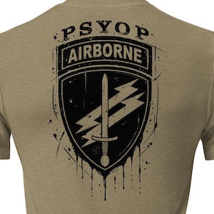 May include: A tan short-sleeve t-shirt featuring a black graphic on the back. The design includes a shield emblem with a sword and lightning bolts, topped with the text PSYOP and AIRBORNE. The graphic has a distressed, dripping ink effect.