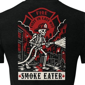 May include: A black short-sleeve t-shirt featuring a graphic on the back of a skeleton firefighter wearing a helmet and holding a hose. The design includes red and white colors, a city background, and the text "FIRE FIRST IN LAST OUT" and "SMOKE EATER".