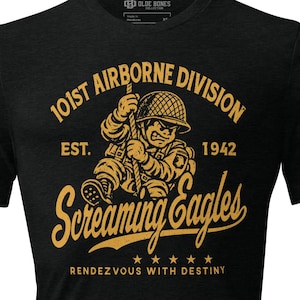 Puede incluir: Una camiseta negra de manga corta con un gráfico dorado de un soldado trepando por una cuerda. El texto dice 101ST AIRBORNE DIVISION, EST. 1942, Screaming Eagles y RENDEZVOUS WITH DESTINY, con cinco estrellas debajo del gráfico principal.