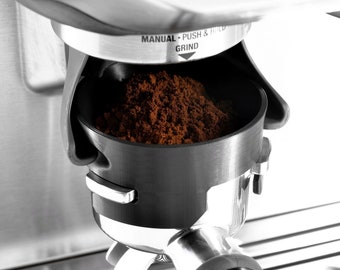 May include: Close-up of a coffee grinder with a black portafilter filled with freshly ground coffee beans. The grinder has a silver finish and a label that reads "MANUAL - PUSH & HOLD GRIND".