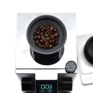 Puede incluir: Un molinillo de caf&eacute; negro con tapa plateada y pantalla digital. El molinillo tiene un dial para ajustar el tama&ntilde;o de la molienda y un peque&ntilde;o taz&oacute;n lleno de granos de caf&eacute;.