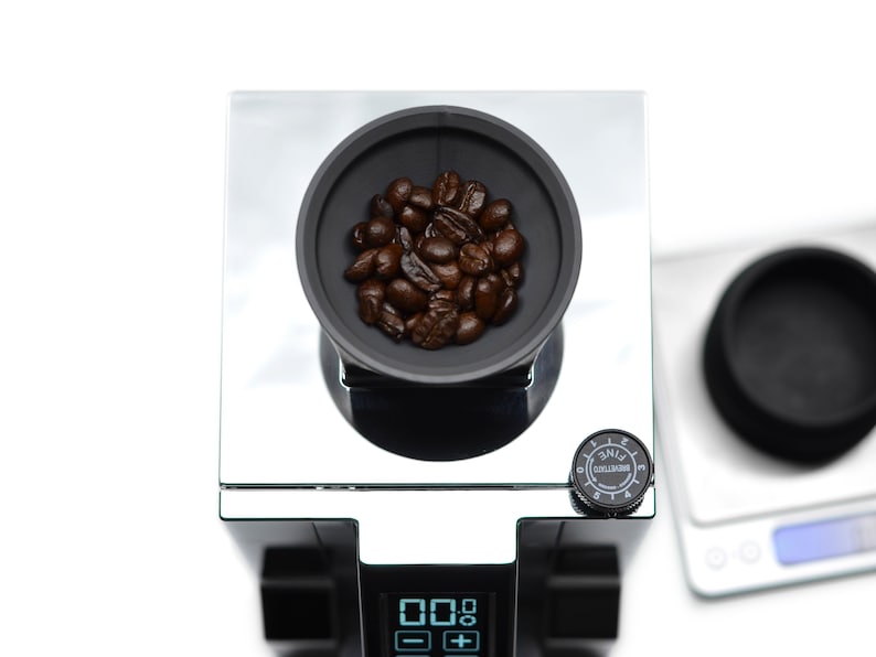 Puede incluir: Un molinillo de caf&eacute; negro con tapa plateada y pantalla digital. El molinillo tiene un dial para ajustar el tama&ntilde;o de la molienda y un peque&ntilde;o taz&oacute;n lleno de granos de caf&eacute;.