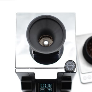 Puede incluir: Un molinillo de caf&eacute; negro y plateado con una pantalla digital y un dial etiquetado como "Grind Setting" con los n&uacute;meros del 1 al 8. El molinillo tiene un dep&oacute;sito negro y una tolva plateada. El molinillo est&aacute; sobre una superficie blanca.