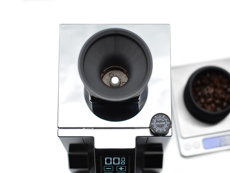 Puede incluir: Un molinillo de caf&eacute; negro y plateado con una pantalla digital y un dial etiquetado como "Grind Setting" con los n&uacute;meros del 1 al 8. El molinillo tiene un dep&oacute;sito negro y una tolva plateada. El molinillo est&aacute; sobre una superficie blanca.
