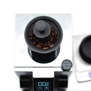 Puede incluir: Una vista de cerca de un molinillo de caf&eacute; negro con tapa plateada. El molinillo tiene una pantalla digital y una perilla con la palabra "SMALL" en ella. El molinillo est&aacute; lleno de granos de caf&eacute;.