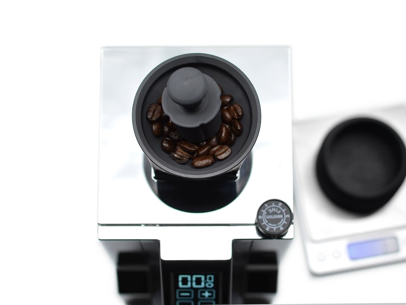 Puede incluir: Una vista de cerca de un molinillo de caf&eacute; negro con tapa plateada. El molinillo tiene una pantalla digital y una perilla con la palabra "SMALL" en ella. El molinillo est&aacute; lleno de granos de caf&eacute;.