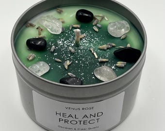 May include: A green candle with black and clear crystals embedded in the wax. The candle is in a silver tin with a label that reads "Venus Rose Heal and Protect Obsidian & Clear Quartz".