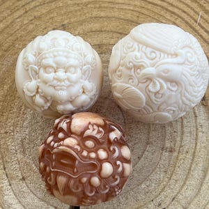 Puede incluir: Tres esferas talladas intrincadamente. Dos son blanquecinas, una que representa una cara y la otra un pájaro. La tercera esfera es marrón y presenta un diseño detallado y texturizado. Estas esferas decorativas son probablemente para exhibición.