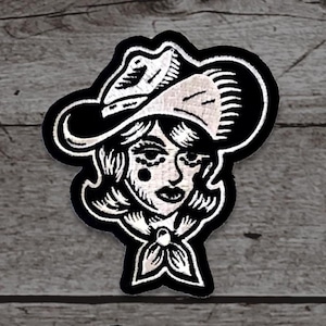 May include: Black and white embroidered patch featuring a cowgirl with a hat, hair, and a neckerchief. The patch has a black border and is designed in a vintage tattoo style. The cowgirl has two beauty marks on her face.