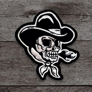 May include: Embroidered patch featuring a skull wearing a cowboy hat and a bandana. The skull is white with black details, and the hat and outline are black. The patch is designed for application on clothing or accessories.