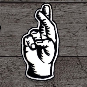 May include: A white patch with a black outline, featuring a hand with crossed fingers. The patch is a symbol of good luck and is set against a gray wooden background. The design is simple and bold.