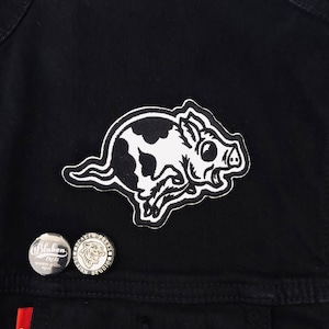 May include: A black and white patch featuring a cartoon boar with a spotted pattern. Two round pins with text and a tiger design are also visible. The patch is sewn onto a black denim jacket.
