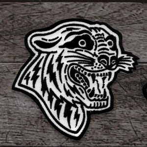 May include: A black and white embroidered patch featuring a roaring tiger head. The tiger's face is detailed with sharp teeth, and lightning bolt-like stripes. The patch is likely for clothing or accessories.