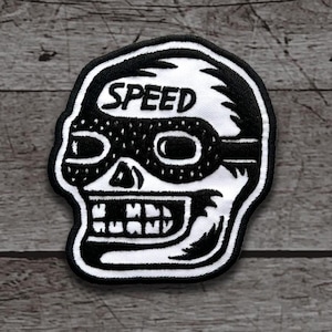 May include: A black and white patch featuring a skull wearing goggles. The word "SPEED" is arched above the goggles. The patch has a black border and is on a gray wooden surface.