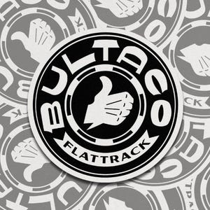 May include: A circular sticker with a black and white design. The sticker features the word "BULTACO" in a bold, sans-serif font, with "FLATTRACK" below. A white thumbs-up graphic is in the center. The background is a pattern of similar stickers.