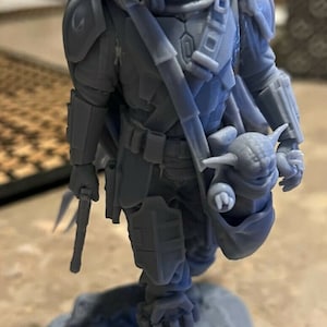 Puede incluir: Una figura gris de un personaje mandaloriano de Star Wars impresa en 3D que sostiene una peque&ntilde;a figura de beb&eacute; Yoda gris. El mandaloriano lleva un casco y una armadura, y sostiene un arma en su mano izquierda.