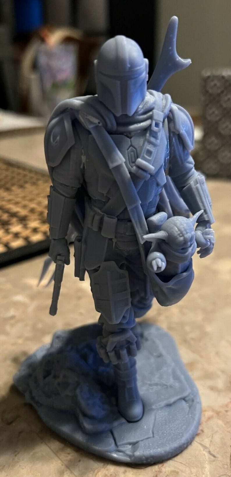 Puede incluir: Una figura gris de un personaje mandaloriano de Star Wars impresa en 3D que sostiene una peque&ntilde;a figura de beb&eacute; Yoda gris. El mandaloriano lleva un casco y una armadura, y sostiene un arma en su mano izquierda.