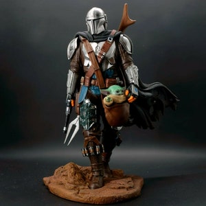 Puede incluir: Una figura de acci&oacute;n de Mandalorian de plata y negro sosteniendo una peque&ntilde;a figura de alien&iacute;gena verde en una bolsa marr&oacute;n. El Mandalorian lleva un casco y una armadura, y el alien&iacute;gena lleva una t&uacute;nica marr&oacute;n. Las figuras est&aacute;n de pie sobre una base marr&oacute;n.