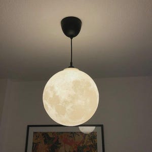 May include: A spherical pendant light, designed to resemble the moon. The light has a textured surface, with a black cord and ceiling fitting. The light is illuminated, creating a soft glow. The fixture is a unique home decor item, with a diameter of approximately 30 cm.
