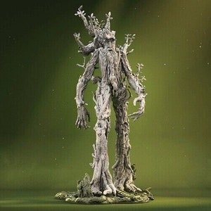 May include: A detailed sculpture of a tree-like humanoid figure with a weathered, bark-like texture. The figure has a human-like form with arms, legs, and a head, but its body is covered in branches and leaves. The sculpture is set on a base of dirt and rocks, suggesting a natural setting.