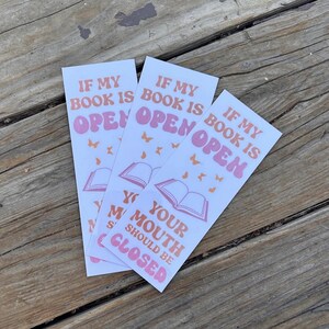 Pode incluir: Três marcadores de livros brancos com o texto "IF MY BOOK IS OPEN, YOUR MOUTH SHOULD BE CLOSED" em rosa e laranja. Os marcadores apresentam ilustrações de livros abertos e borboletas.