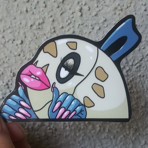 Könnte beinhalten: Ein weißer Aufkleber mit einer Comicfigur mit blauen und rosa Akzenten. Die Figur hat große Augen, rosa Lippen und lange Wimpern. Die Figur trägt blaue Handschuhe mit rosa Nägeln.