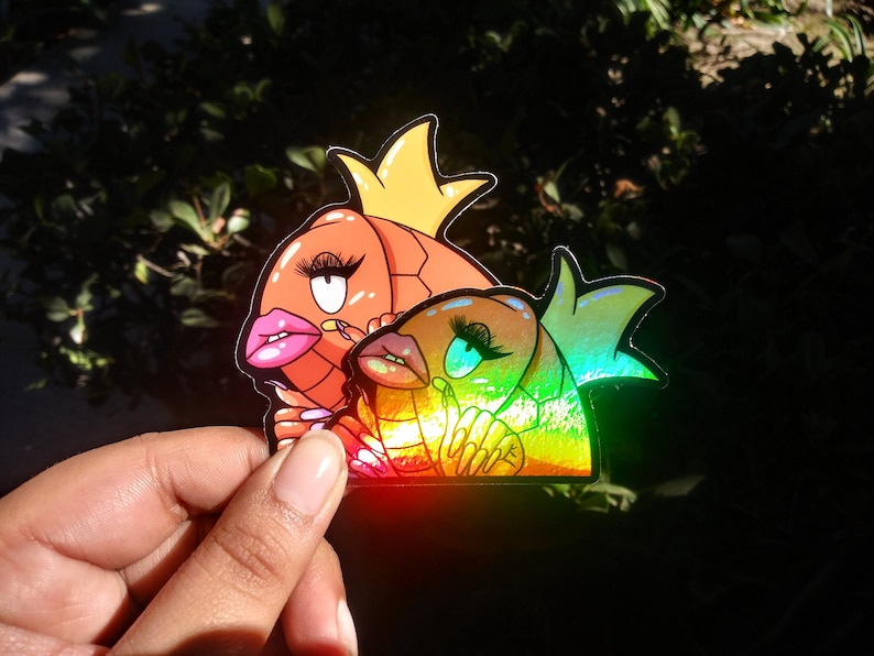 Puede incluir: Dos pegatinas hologr&aacute;ficas iridiscentes de peces de dibujos animados con ojos grandes y pesta&ntilde;as. Los peces llevan coronas y tienen labios rosados. Las pegatinas est&aacute;n sobre un fondo oscuro.
