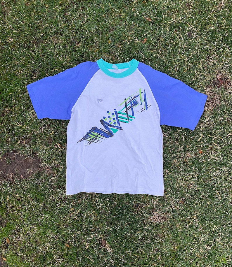 May include: White t-shirt with a blue and green abstract design. The shirt has a blue and turquoise raglan sleeve.