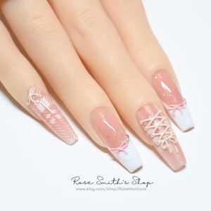 May include: A hand with long, square-shaped artificial nails. The nails feature a pink and white design with intricate lace-up and bow details. The base of the nails is a soft pink, transitioning to white tips. The text "Rose Smith's Shop" is visible.